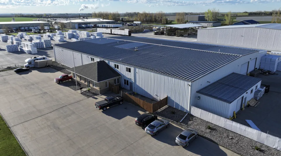 Aerial view of a large industrial warehouse with parked cars and stacks of pallets in the background under a blue sky.