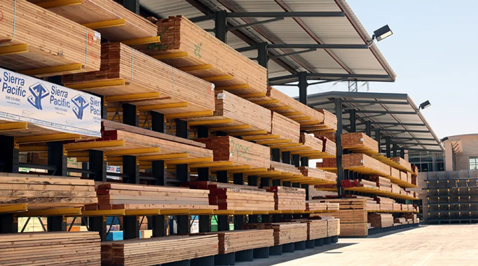 Outdoor lumber yard with stacked wooden planks under metal roofing and clear sky above