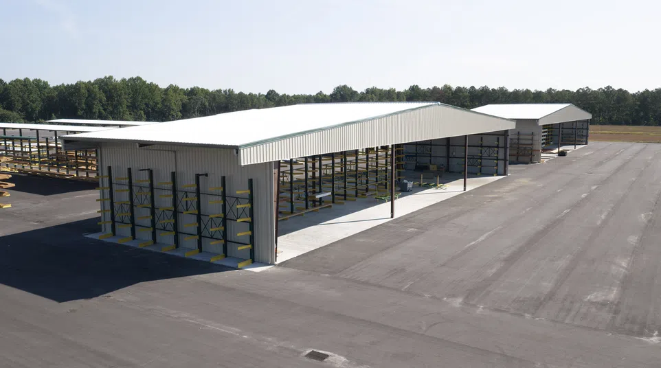 Two large metal storage sheds with open sides and cantilever rack shelving on a wide paved lot under clear sky