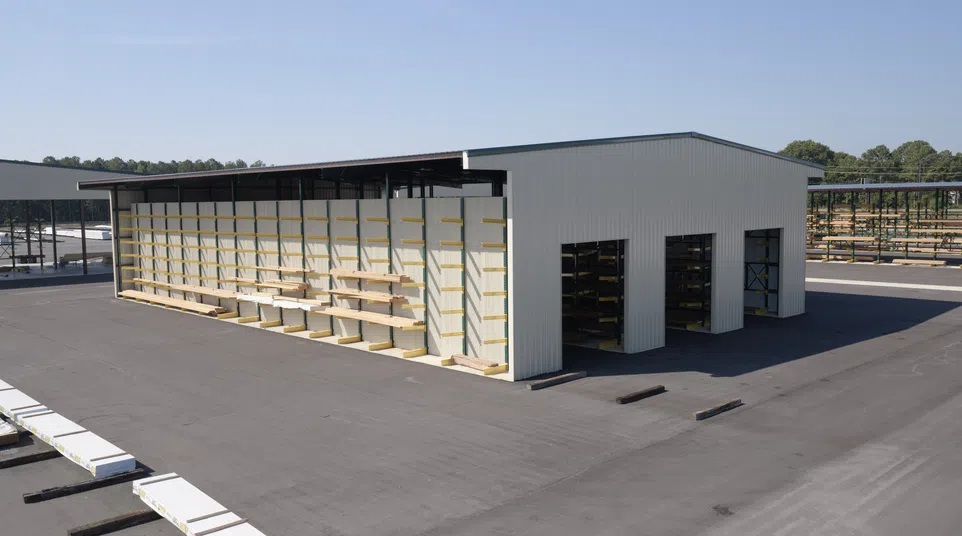 Large outdoor storage facility with metal building and wood planks stored on exterior racks under clear sky