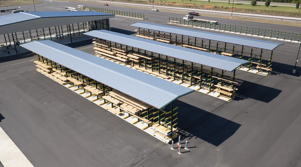 Large outdoor lumber storage racks with metal roofs in a paved yard beside a road and greenery in the background