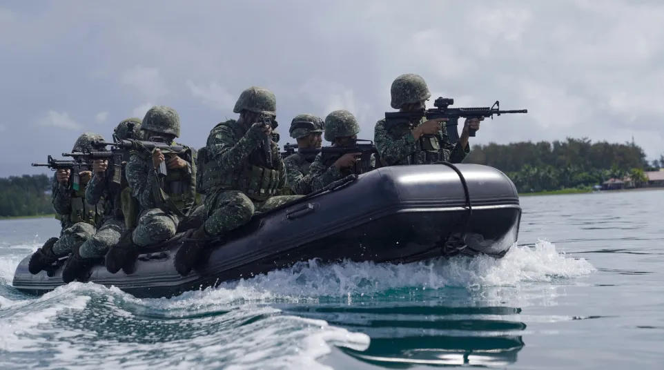 Soldiers in camouflage gear aiming rifles while riding a black inflatable boat on water during training.