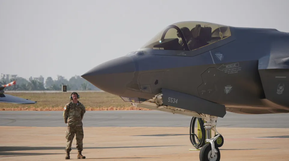 Military personnel standing next to a parked stealth fighter jet on an airfield during daytime