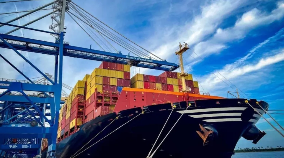 Large cargo ship loaded with colorful containers docked at port under blue sky with cranes in background