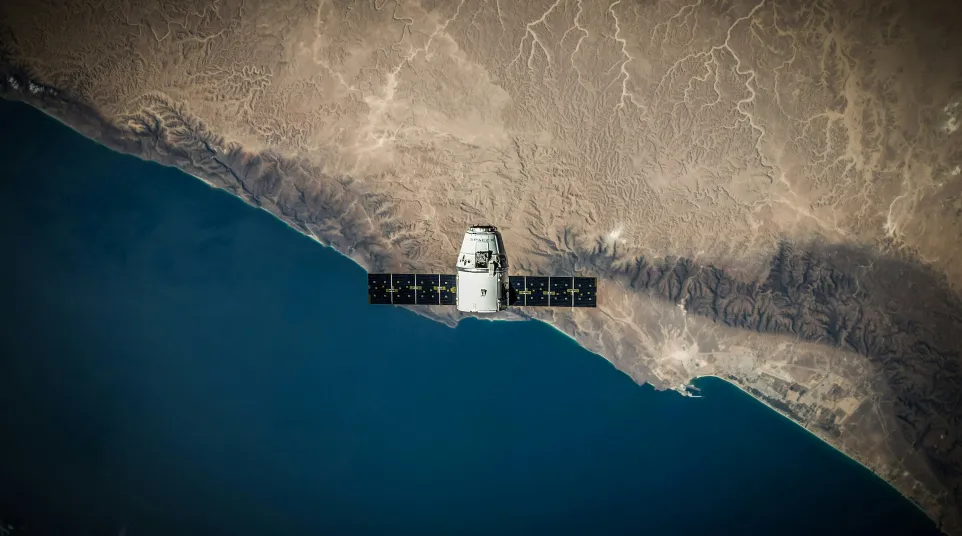Satellite with solar panels orbiting over coastal desert and deep blue ocean beneath clear sky.