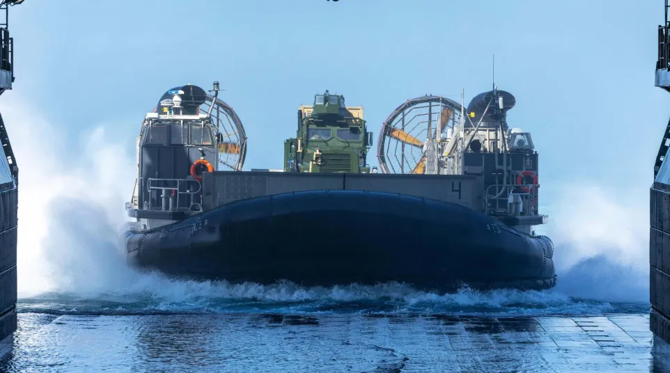 Military hovercraft launching from ship ramp creating water spray under blue sky.