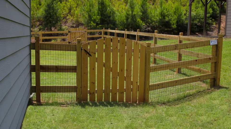 Wooden fence with wire mesh surrounding a grassy backyard area next to a house and trees.