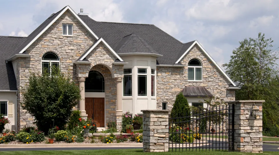 Elegant stone house with a landscaped garden and decorative gate under a bright sky.