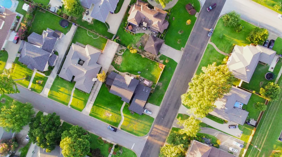 Aerial view of suburban neighborhood with houses, green lawns, and winding streets under a bright sky.