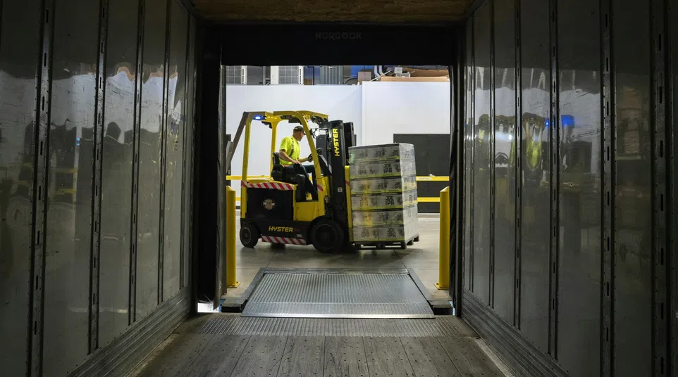 Forklift operator loading stacked pallets into a trailer through a warehouse loading dock with metal walls.