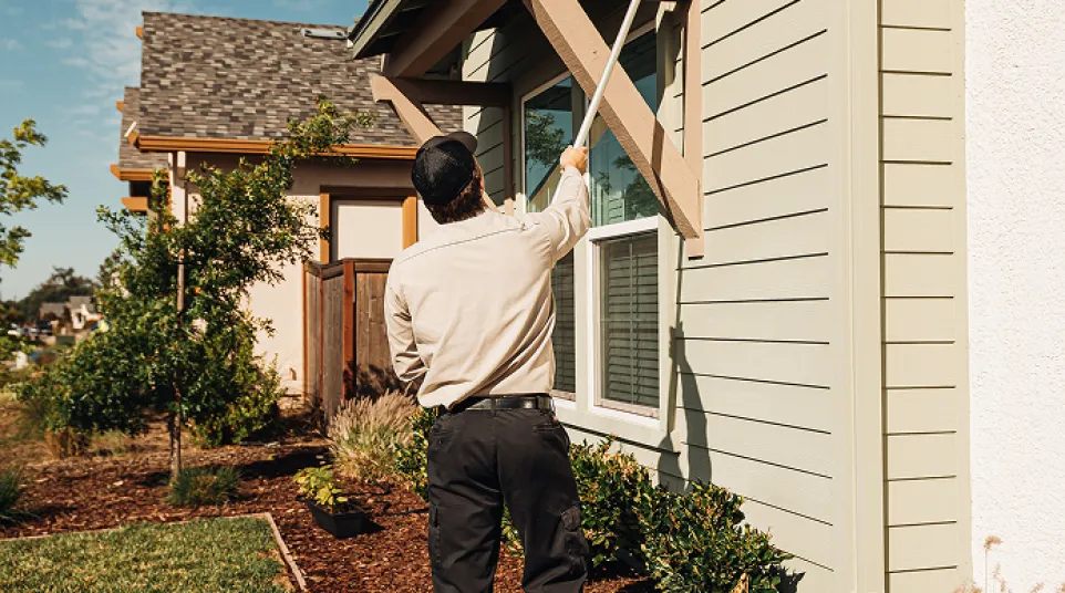 Man cleaning exterior of house with long-handled duster on sunny day in suburban neighborhood