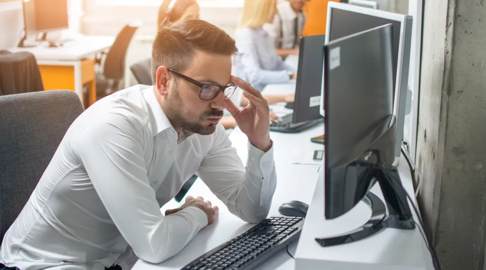 Frustrated young professional working at a computer in an office environment with colleagues in the background.
