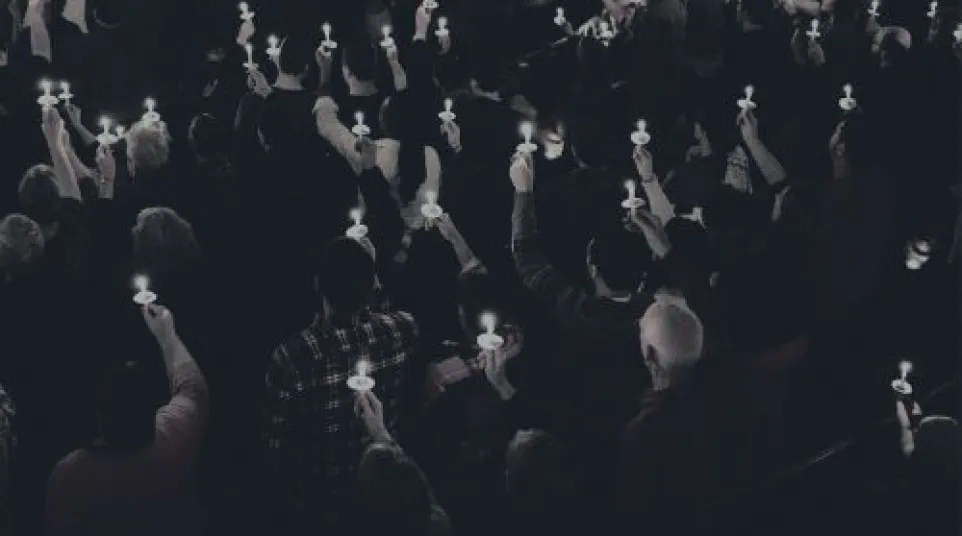Large crowd holding lit candles in a dark setting, forming a solemn vigil or memorial gathering.