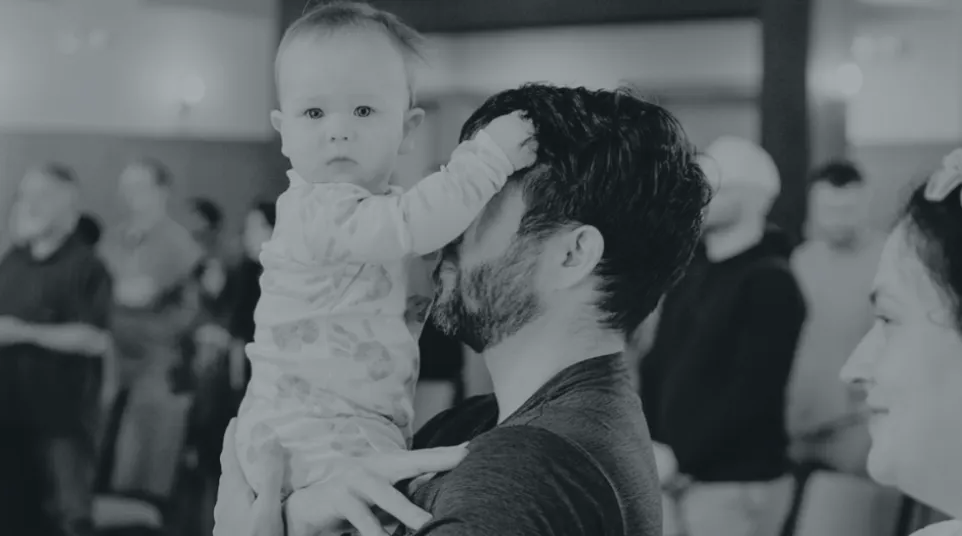 Father holding baby who touches his face in a gathering with seated and standing people in the background.