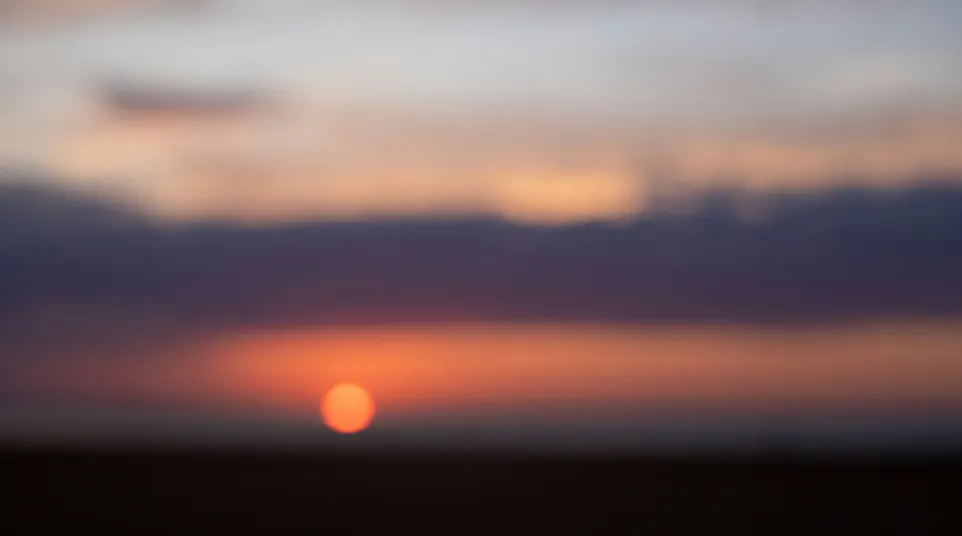Blurred sunset with orange sun and dark clouds over a flat horizon during dusk.