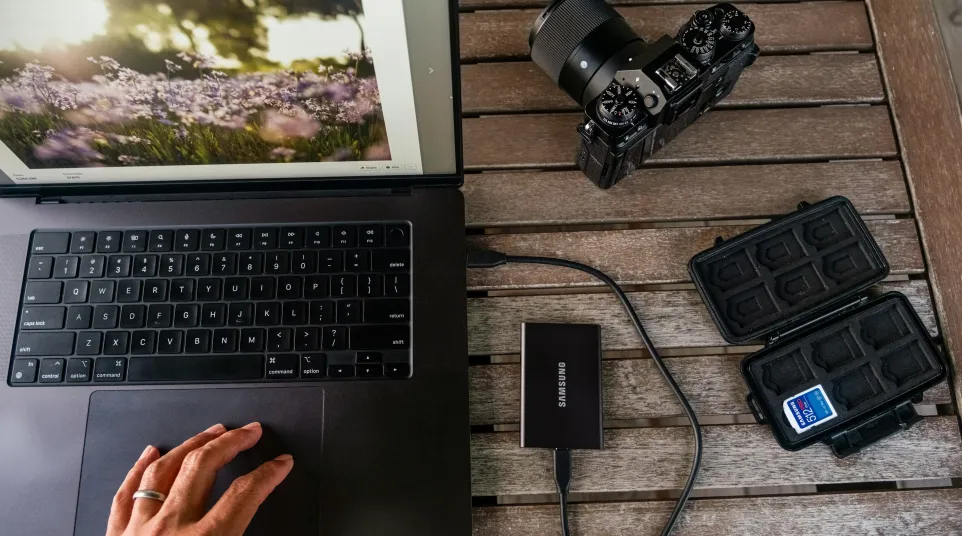 Hand using laptop next to a camera, external Samsung SSD, and SD card case on wooden table.