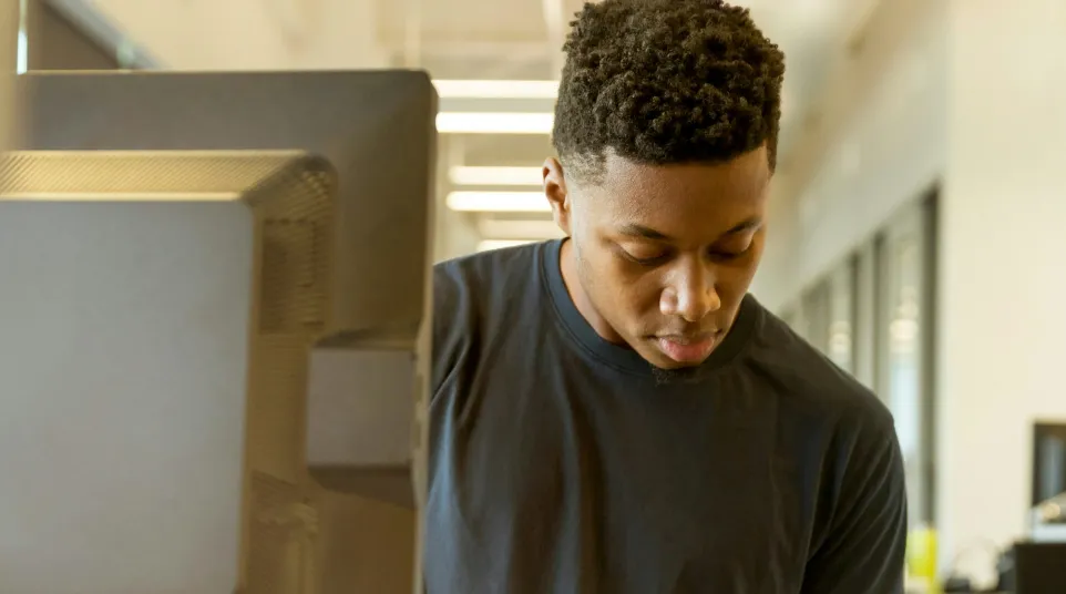 Young man standing at desk in office, writing notes in notebook with computer and keyboard nearby