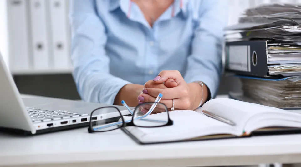 Person sitting at desk with laptop, open planner, eyeglasses, and large stack of paperwork in office