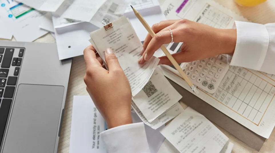 Person organizing and reviewing receipts with pencil, calculator, laptop, and paperwork on a desk for budgeting.