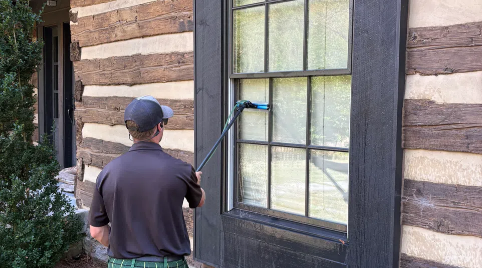 Man in a green plaid kilt cleaning a large window on a rustic log cabin with a window cleaning tool.