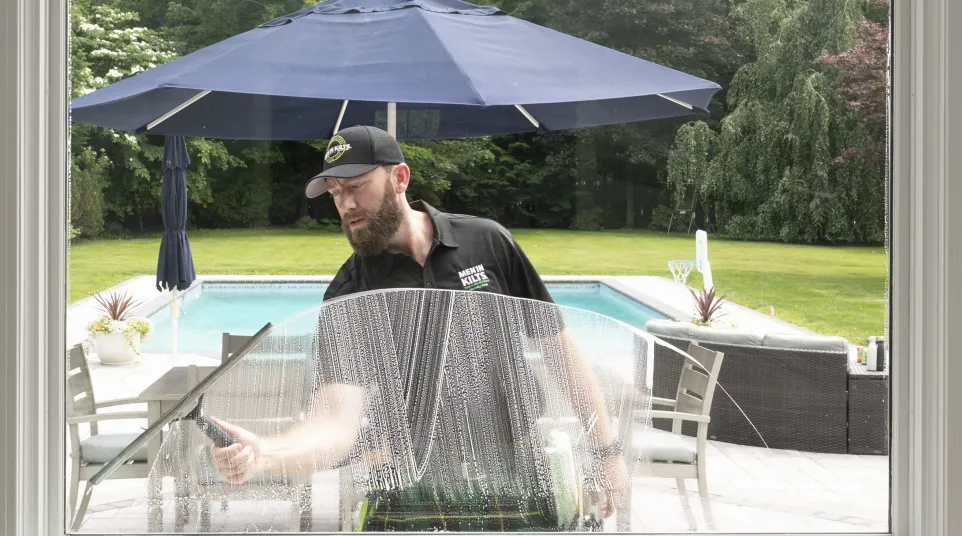 Man cleaning a window with a squeegee outside near a pool and patio with blue umbrella and greenery.