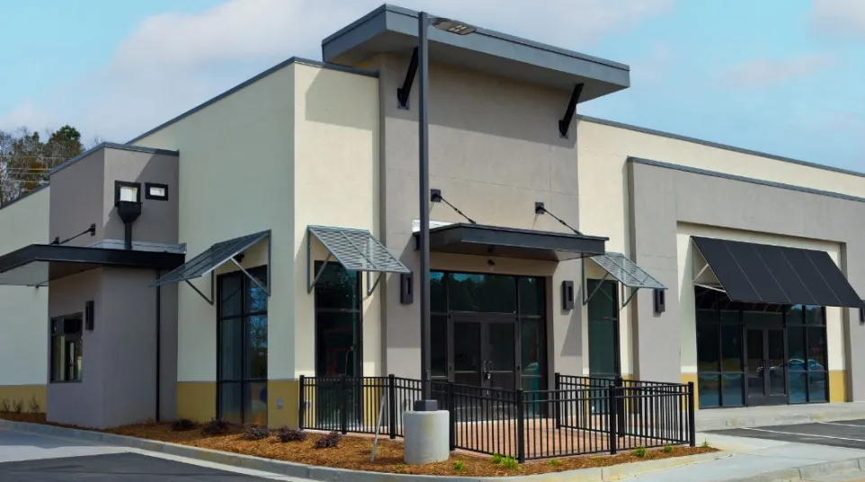 Modern commercial building with large windows, metal awnings, beige and gray exterior, and black railing on a sunny day.