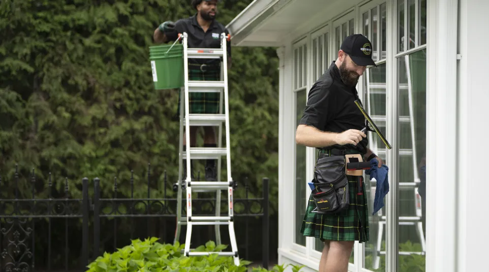 Two window cleaners in kilts cleaning large glass windows outside a building with greenery around.