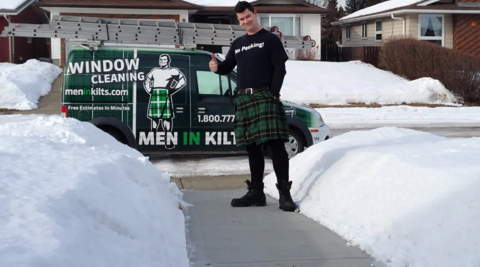 Man wearing a green tartan kilt stands on snow-lined sidewalk giving thumbs up next to a Men In Kilts window cleaning van.