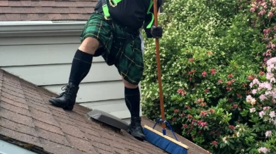 Man wearing a green kilt and safety harness sweeping moss off a sloped roof with a blue brush near flowering bushes.