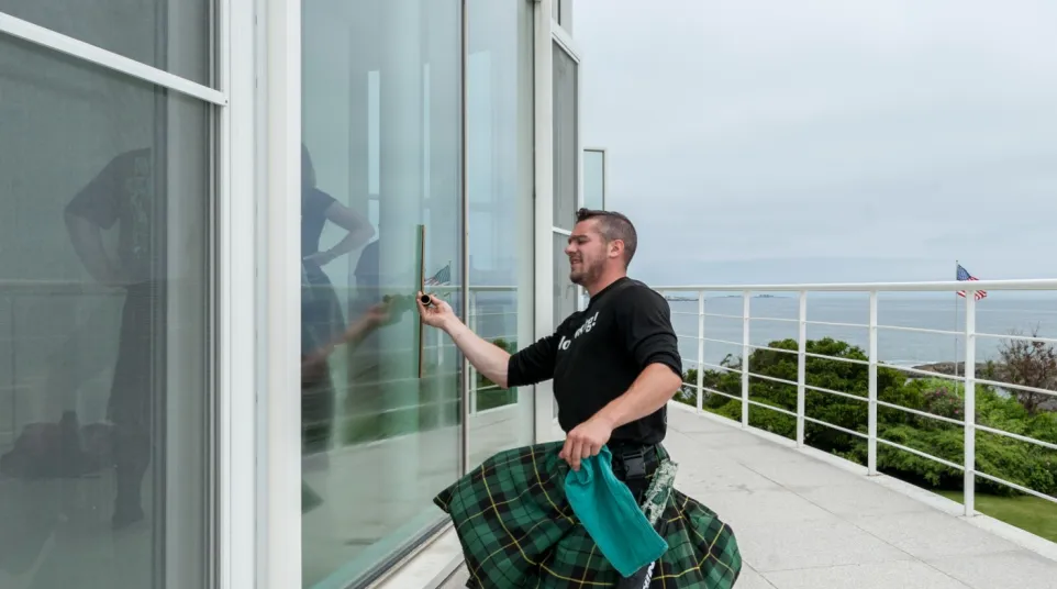 Man wearing a green kilt cleans large glass windows on a modern balcony overlooking the ocean.