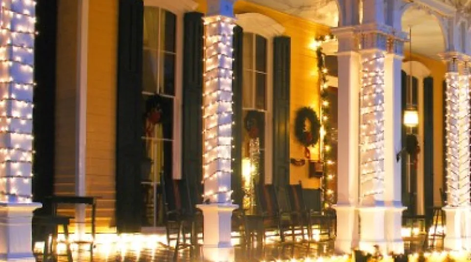 Large porch of a yellow house decorated with white Christmas lights wrapped around columns and railings at dusk.