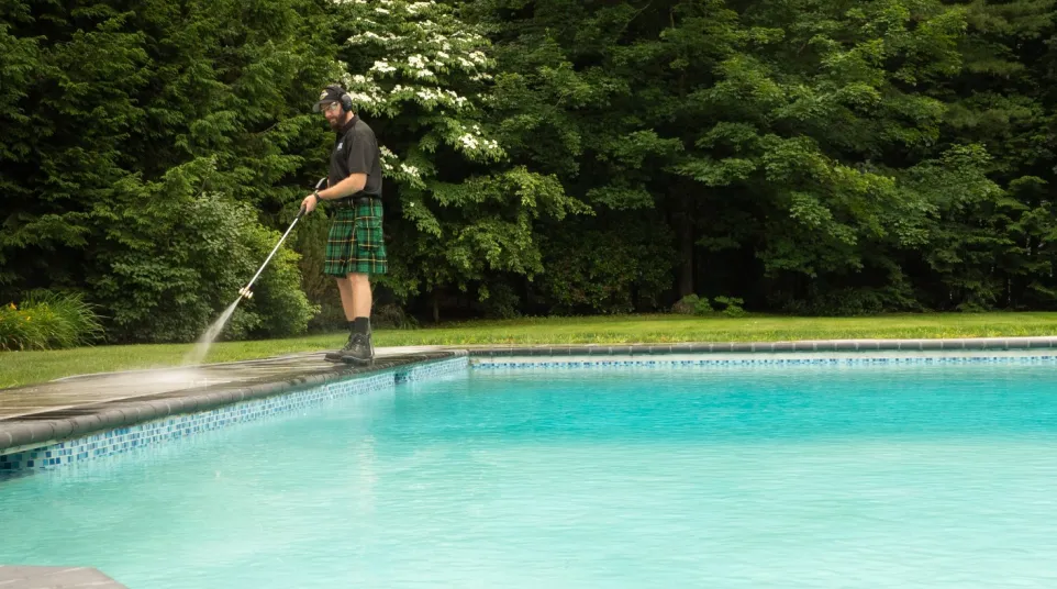 Man wearing a kilt pressure washing the edge of a backyard swimming pool surrounded by green trees.