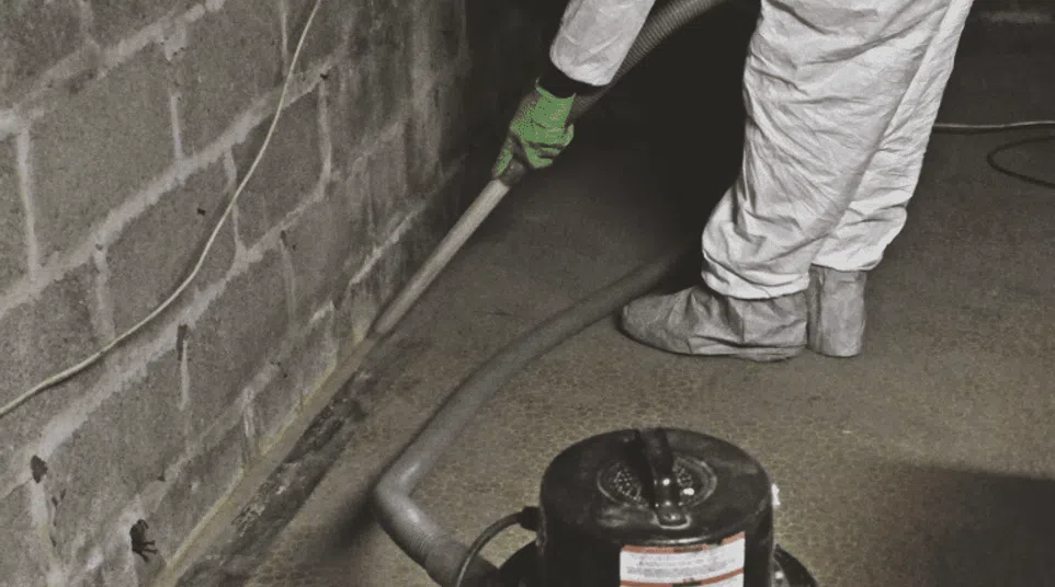 Person in protective suit and gloves vacuuming dust and debris from basement floor near concrete wall.