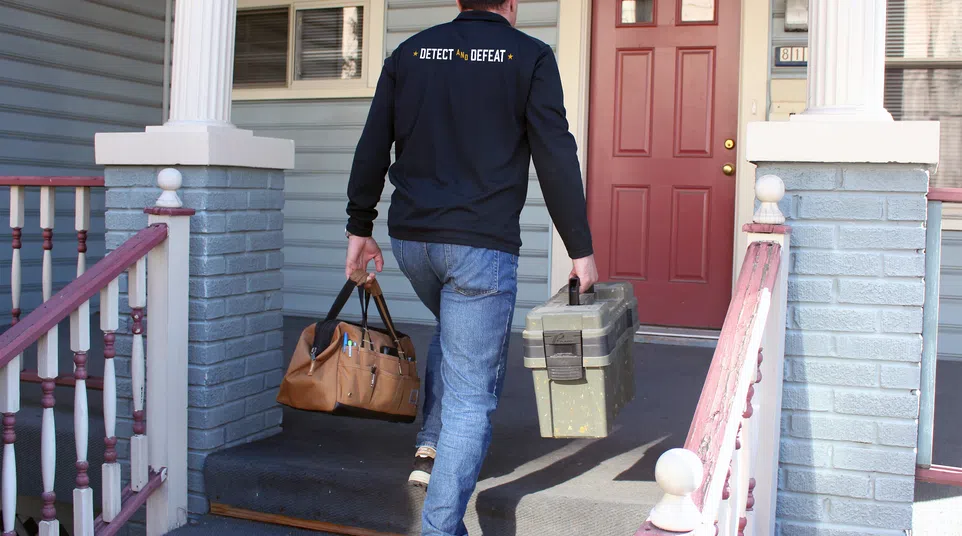 Home inspector carrying tool bags up porch steps to inspect house with red door and gray siding.