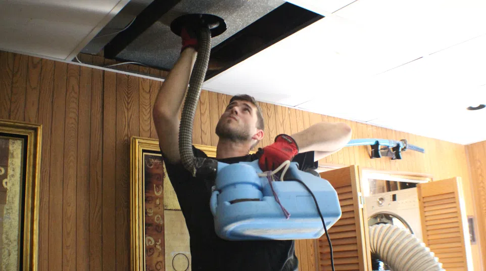 Technician using a blue machine to clean a ceiling vent in a wood-paneled room wearing red gloves.