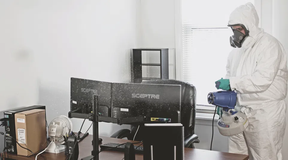 Person in protective suit disinfecting office workspace with computer monitors and desk fan.
