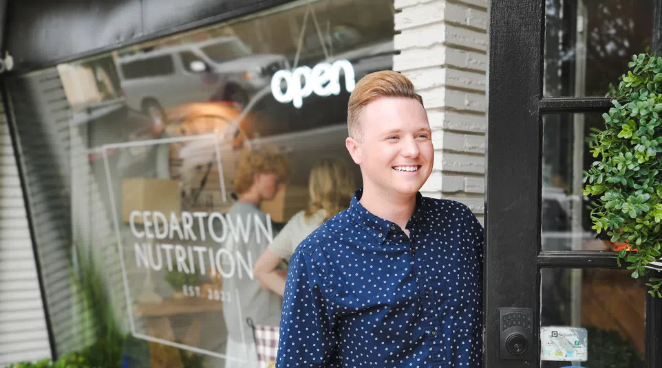 Smiling man stands at the entrance of Cedartown Nutrition store with open sign and greenery outside