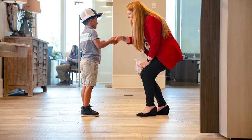 Woman in red jacket smiling and shaking hands with young boy wearing a cap in a bright room