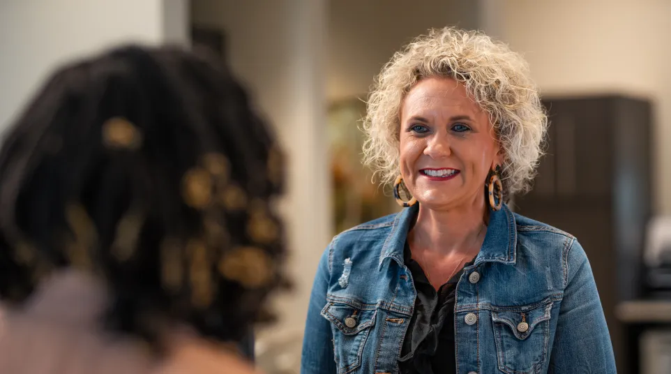 Smiling woman with curly hair in denim jacket talking to another person indoors in a casual setting