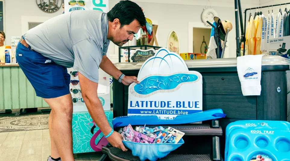 Man arranging Latitude.Blue branded pool accessories and sunglasses inside a retail store near hot tub display.