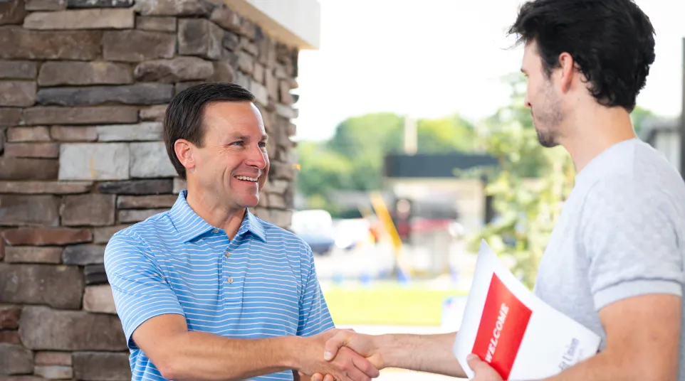 Two men shaking hands outside a building, one holding a welcome brochure, smiling and greeting.