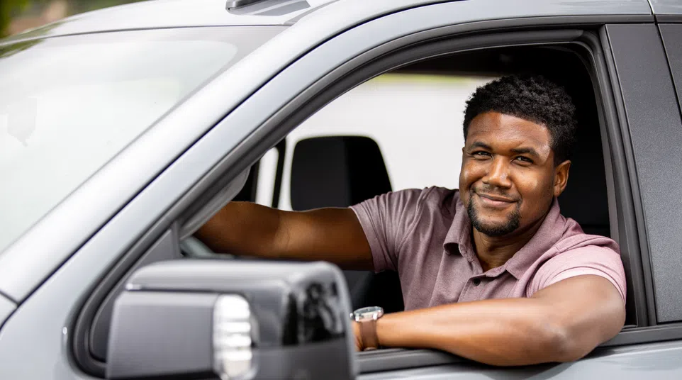 Smiling man in a purple shirt sitting in the driver's seat of a gray car with his arm resting on the window.