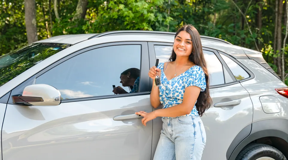 Smiling young woman holding car keys while standing next to a silver SUV with trees in background