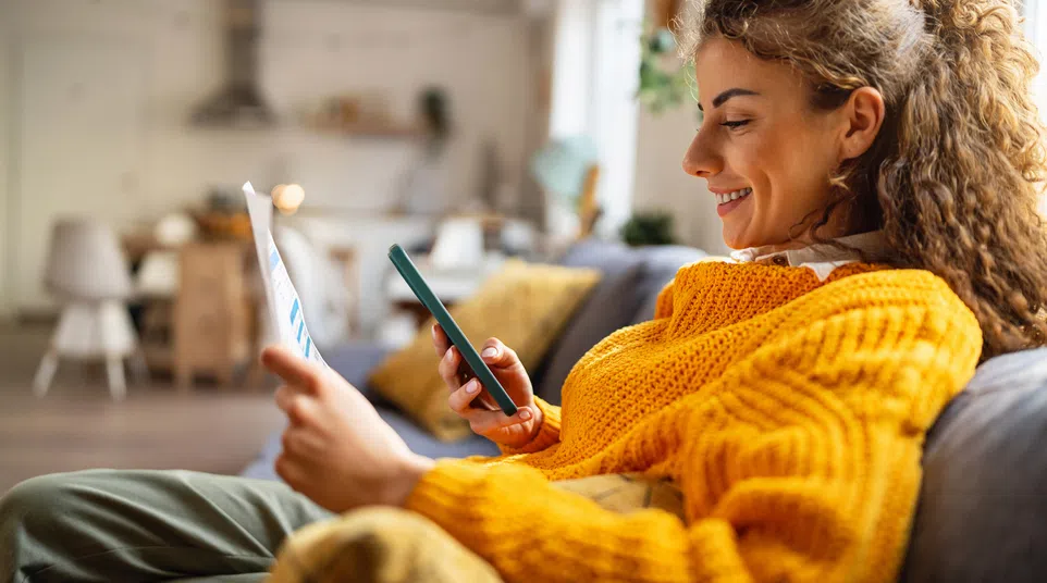 Smiling woman in a mustard sweater using smartphone while holding paper on a cozy living room couch.