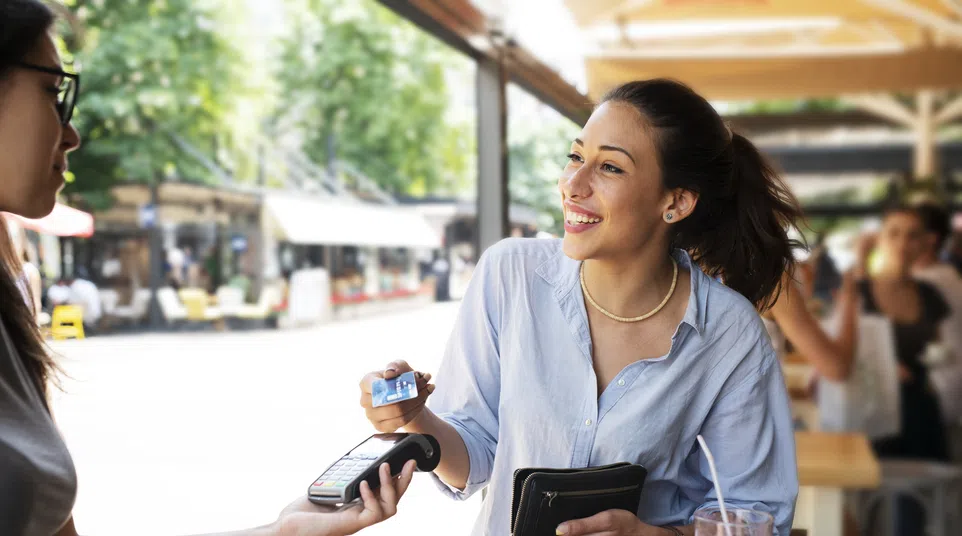 Smiling woman paying with credit card at outdoor café while holding wallet, with drinks on the table.