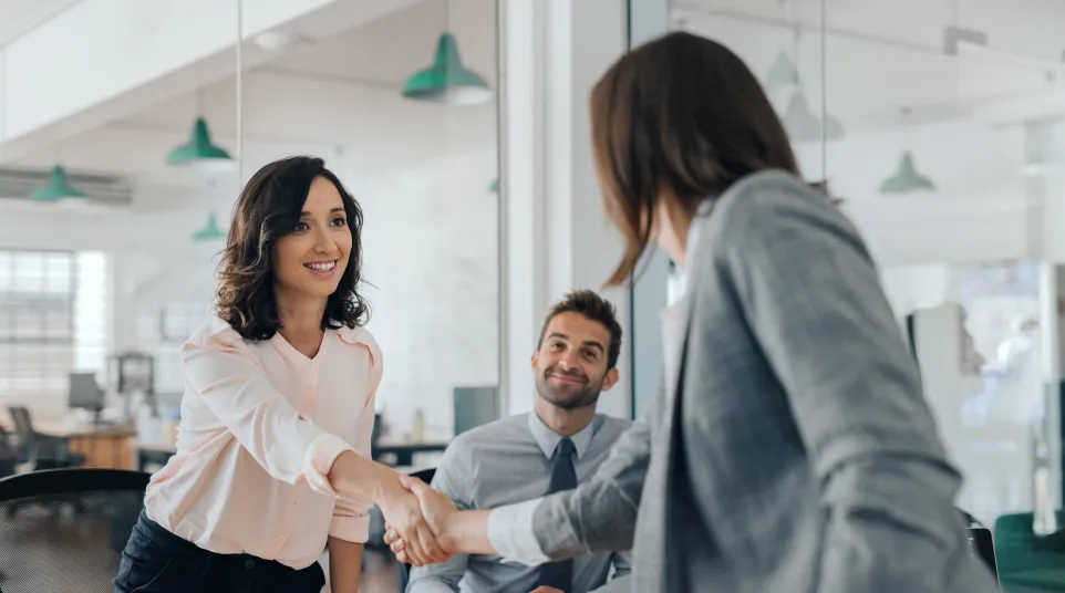 Two businesswomen shaking hands during a meeting with a man smiling in a modern office setting