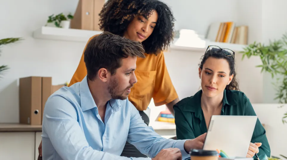 Three colleagues collaborate on a laptop in a modern office with plants and shelves in the background