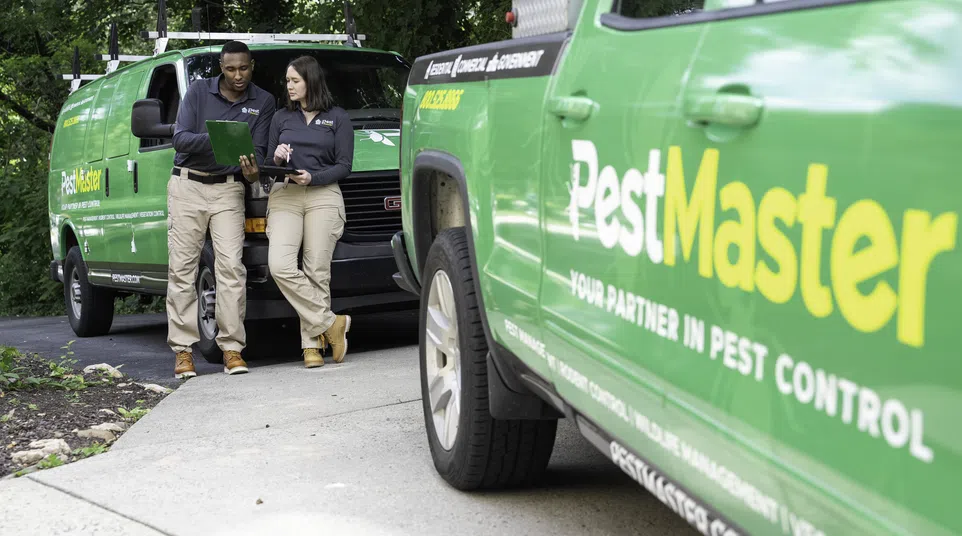 Two PestMaster workers in uniform review a tablet near green branded pest control vehicles on a driveway.