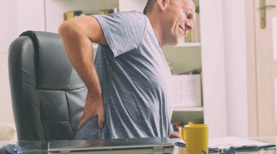 Middle-aged man sitting in office chair holding lower back in pain expressing discomfort indoors.