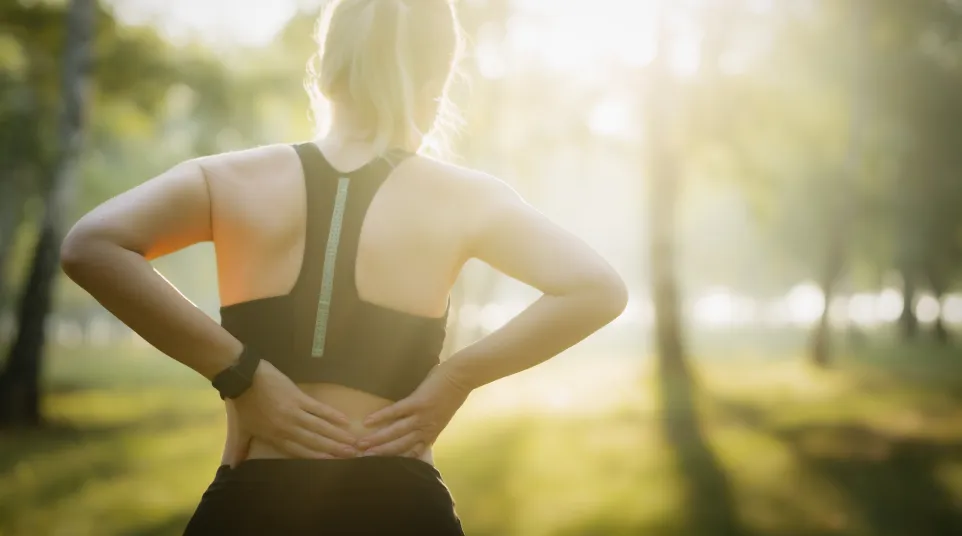 Woman in sportswear holding lower back outdoors in a sunlit park, indicating back pain or discomfort.
